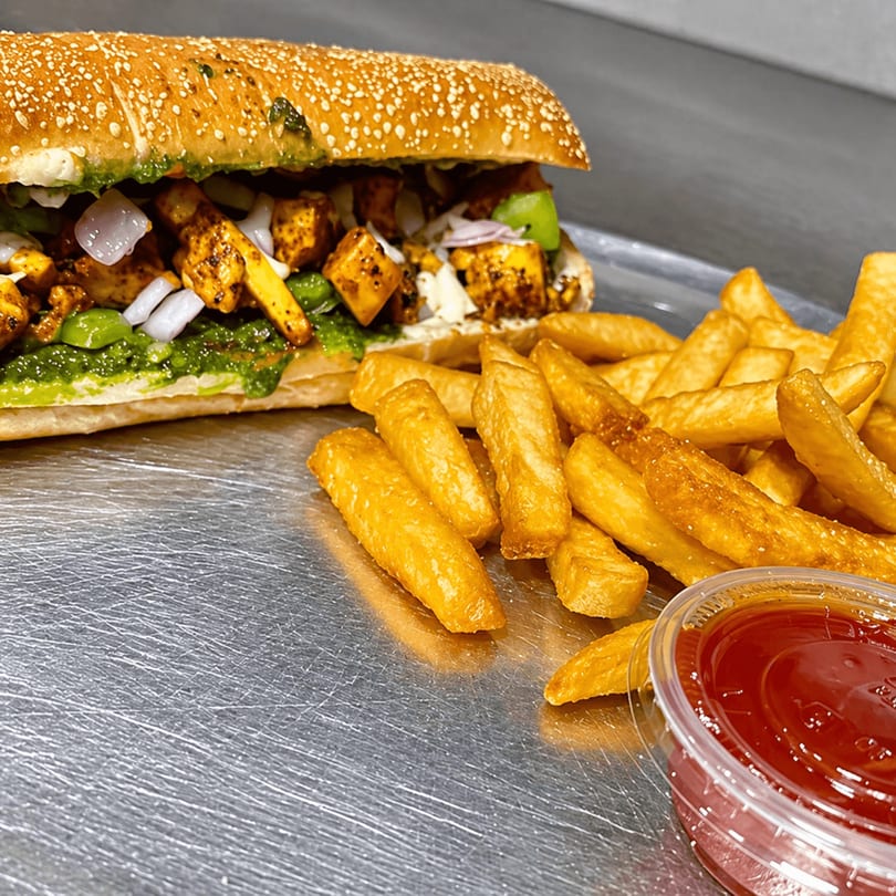 A paneer tikka sub sandwich with green chutney, onions, and bell peppers, served with fries and ketchup.